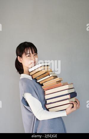Portrait of puzzled Asian student girl in knitted vest carrying big stack of books against isolated background Stock Photo