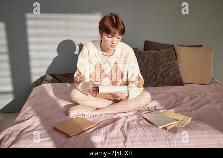 Serious redhead student girl in casual sweater sitting with crossed legs on bed among books and examining notes in sketchpad Stock Photo