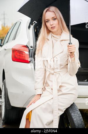 Vertical portrait of beautiful woman on road near her stopped white car with punctured car tire. Female driver waiting for help, holding umbrella, sitting on spare wheel. Stock Photo
