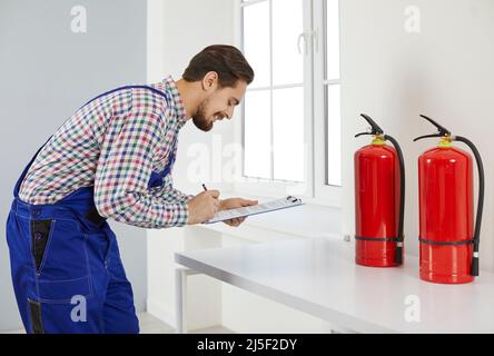 Happy fire safety engineer writing on clipboard while checking condition of fire extinguishers Stock Photo