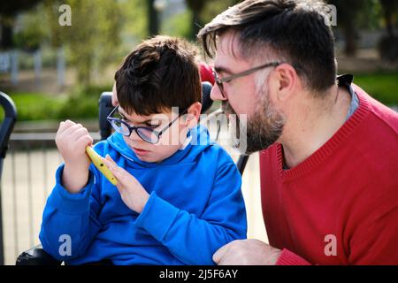 Disabled child on wheelchair using a mobile phone with his father. Stock Photo