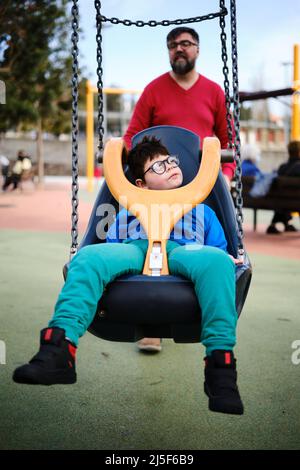 Disabled boy swinging on a special needs swing while enjoying with his father at the playground. Stock Photo