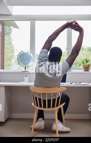 Rear View Of Man Working From Home On Computer  In Home Office Stretching At Desk Stock Photo