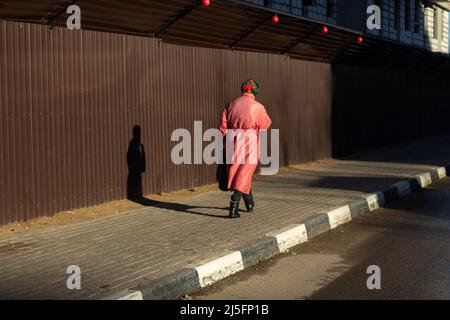 A Russian grandmother walks down the street. An old woman in Russia. Pensioner in a pink cloak. A man walks along the fence. Stock Photo