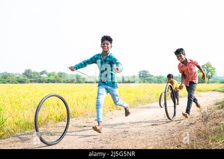 happy cheerful Indian village kids playing with tyre wheel rolling near paddy field rural street - concept of entertainment, holidays and leisure Stock Photo