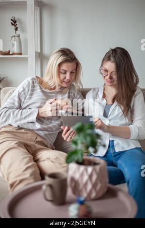 Two young women friends using tablet together, sitting on the couch and smiling Stock Photo
