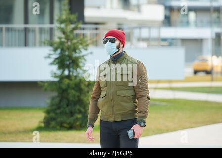A man in a medical face mask holding his smartphone and walking down the street. Stock Photo