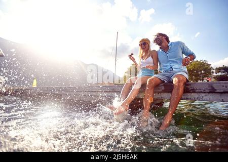 Attractive couple laughing while sitting on jetty near water. Splashing water with legs. Tourism, summertime, togetherness, lifestyle concept. Stock Photo