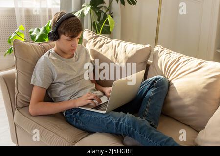 Portrait of teenager boy lying on a sofa in a room, wearing black headphones , looks into a gray laptop Stock Photo