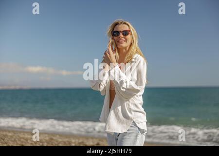 Active lady in sunglasses smiling while calling someone, standing on a beach Stock Photo