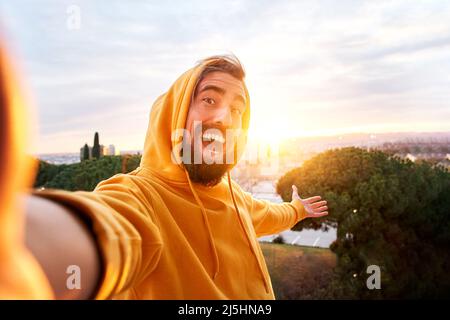 Selfie of young smiling man hooded sweatshirt. Happy Guy with good laugh taking self picture at sunset showing beautiful views. Cheerful traveler Stock Photo