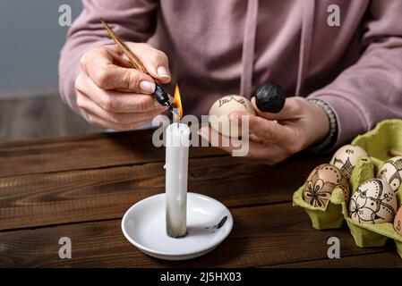 Female hands traditionally paint eggs with floral patterns using a fountain pen and wax for Easter on wooden table. Easter preparation concept. Stock Photo