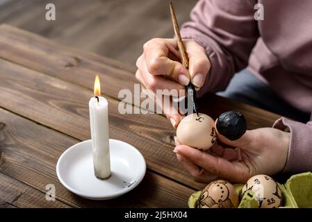 Female hands traditionally paint eggs with floral patterns using a fountain pen and wax for Easter on wooden table. Easter preparation concept. Stock Photo