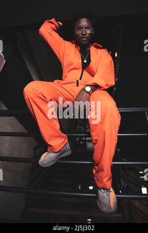 Black prisoner in an orange jumpsuit sitting on his cell fence, posing. Stock Photo