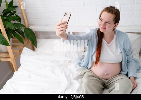 Beautiful cheerful pregnant woman taking selfie photo using mobile phone lying on bed at home. big belly advanced pregnancy. Stock Photo