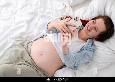 Beautiful cheerful pregnant woman taking selfie photo using mobile phone lying on bed at home. big belly advanced pregnancy. Stock Photo