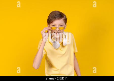 Studio happy smiling young male wearing bright t-shirt and round glasses, with headphones around his neck, listening to his favorite music, looking in on yellow background. Caucasian teenager posing Stock Photo