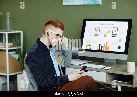 Side view portrait of trendy young businessman using smartphone while working at desk in green office Stock Photo