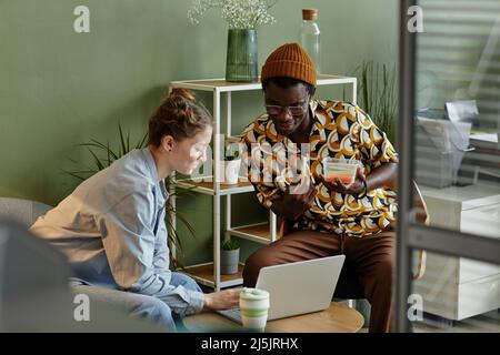 Portrait of two young people using laptop while relaxing in office lounge behind glass wall Stock Photo