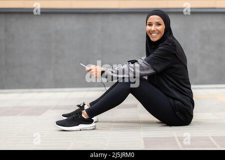 Glad young islamic woman athlete in hijab, sportswear with smartphone sits on floor on gray wall background