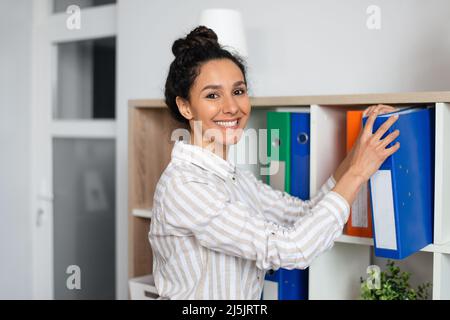 Cheerful female financier doing paperwork, taking folders from shelf, looking and smiling at camera, working in office Stock Photo
