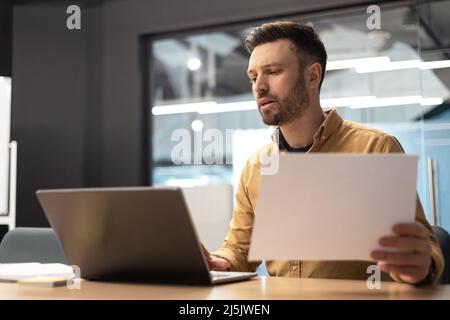 Businessman Using Laptop Holding Paper Working At Workplace In Office Stock Photo
