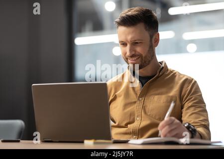 Cheerful Entrepreneur Using Laptop And Taking Notes Working At Workplace Stock Photo