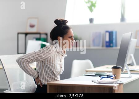 Young businesswoman having acute back pain, massaging aching muscles, sitting at desk in office interior, copy space Stock Photo