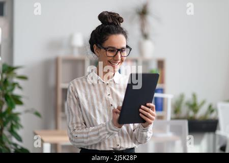 Positive young female entrepreneur using digital tablet in office and smiling, checking her agenda on pad Stock Photo