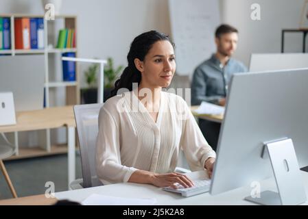 Focused young woman working on computer at open space office with her male colleague Stock Photo
