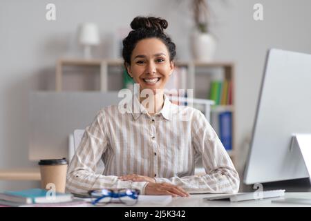 Portrait of cheerful female manager sitting at workplace at contemporary office and smiling at camera Stock Photo