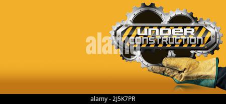 Gloved hand holding an under construction sign with metal cogwheels (gears), on orange and yellow background with copy space and reflections.