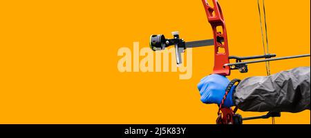 Close-up of the hand with blue glove of an archer aiming with his red hunting compound bow, isolated on an orange background with copy space. Stock Photo