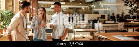 Nice talk. Three young colleagues in casual wear holding coffee cups and discussing something while standing in the modern office Stock Photo