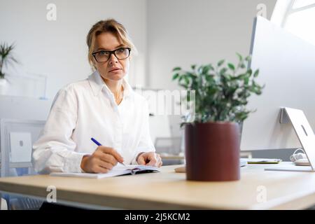 Mature woman office worker writes in a notebook while sitting in the workplace Stock Photo