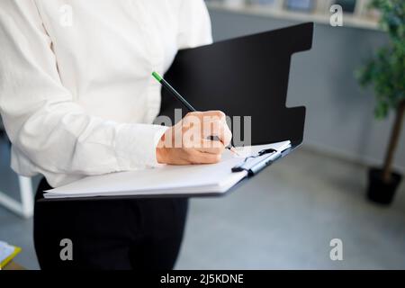 Close up view of female office worker writes on documents using pencil and clipboard Stock Photo