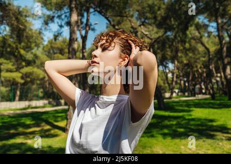 Calm beautiful young woman wearing white tees styling her hair while enjoying fresh air outdoor, relaxing looking away. Feeling alive, breathing, drea Stock Photo