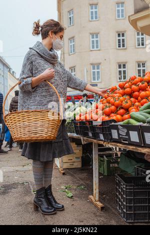 Buying fresh bio vegetables at the grocery market. girl with wicker basket for groceries chooses ripe tomatoes on counter of grocery market. Stock Photo