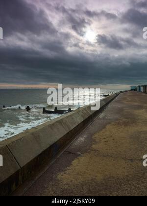 A sweep of sea wall and promenade at Frinton, standing against breaking waves under a stormy sky with a tear admitting sunlight. Stock Photo