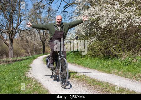 Funny senior man rides his bike hands-free on a dirt road, past flowering sloe bushes and laughs heartily at the camera. He enjoys cycling into spring. Stock Photo