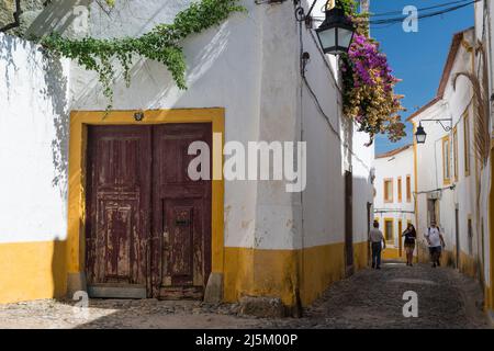 Three persons walking in one of the picturesque cobble stoned alleys with whitewashed and yellow houses in the historical center of Evora, Portugal. Stock Photo