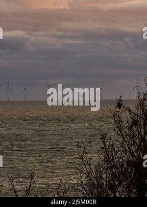 The silhouetted turbines of a wind farm, guarded by the hulk of a Maunsell Fort, stand on the horizon in the North Sea under a dramatic, stormy sky. Stock Photo