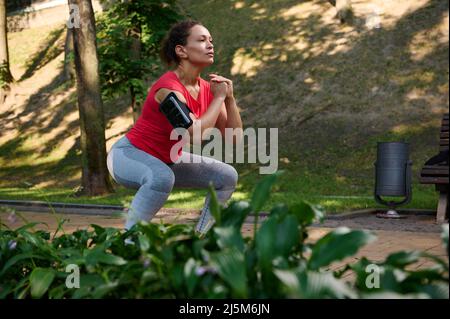 Determined athlete, sportswoman exercising outdoor, doing deep squats in the city park. Enjoy summer days and keep your body fit. Sport, fitness, func Stock Photo