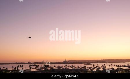 Palm tree silhouettes by ocean harbor at sunset, San Diego, California coast, USA. Coronado island and yacht boats in harbour, palmtrees by marina in bay orange sky. Naval helicopter flying in air. Stock Photo