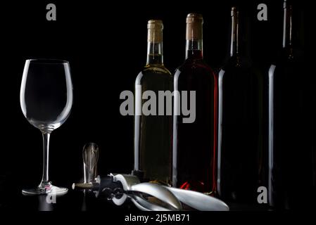 Several bottles of red, rose and white wine in a row, next to an empty glass and a corkscrew on the table. Stock Photo