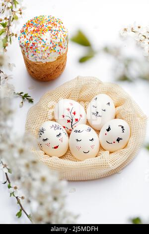 White Easter eggs in a mesh eco-bag with beautiful painted faces and delicious Easter on a white background. Fresh flowering cherry twigs. Vertical photo. Happy Easter. Stock Photo