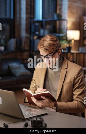 Concentrated young manager making notes in diary and viewing report while working late in office Stock Photo