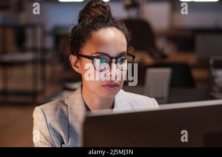 Concentrated young mixed race businesswoman with hairbun wearing eyeglasses looking at computer monitor in dark office Stock Photo