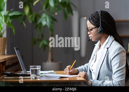 Focused african female home-based virtual assistant in headset making notes during online meeting Stock Photo