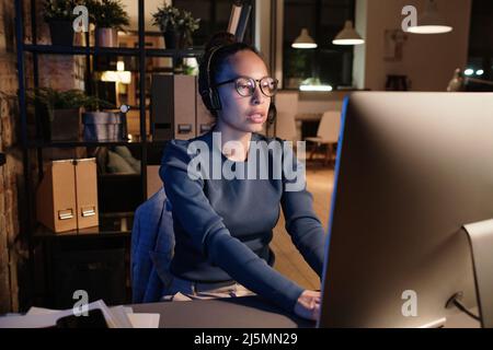 Female call center operator in headset with microphone typing on computer and talking to customer while working night shift Stock Photo